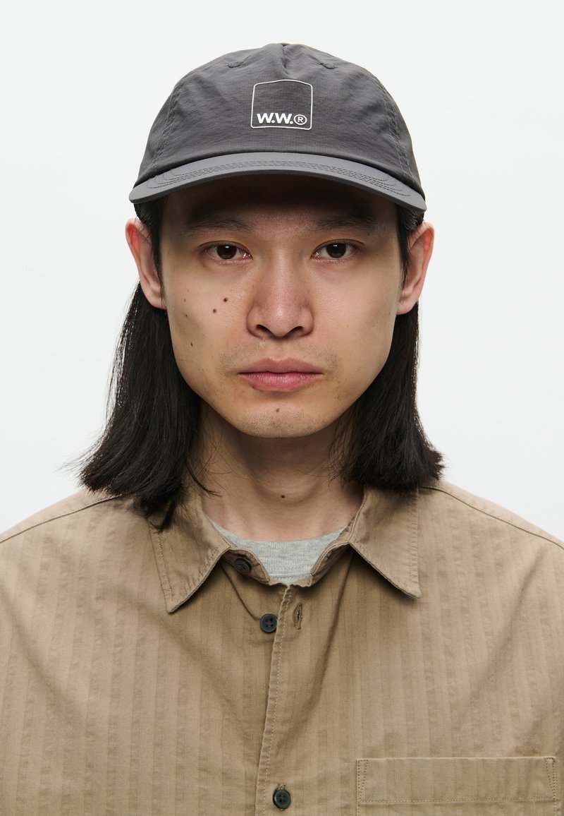 Uomo asiatico con capelli neri lunghi fino alle spalle, che indossa un berretto grigio con il logo "W.W.®" e una camicia button-up marrone chiaro sopra una maglietta grigia, rivolto verso l'avanti.