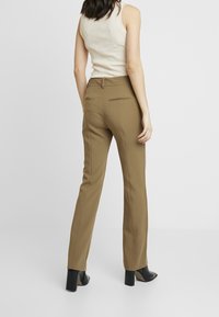Pantalons marron sur mesure avec une texture lisse, coupe ajustée, poches passepoilées à l'arrière, et associés à des chaussures à talons noires. Design simple et fonctionnel.