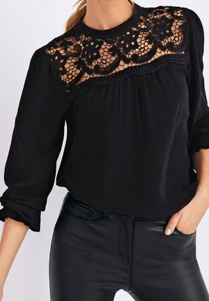 Blouse noire avec un panneau en dentelle aux épaules, poignets froncés et coupe décontractée. Associée à un pantalon noir ajusté pour un look élégant.