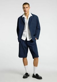 Marineblå letvægtsjakke med en enkelt brystlomme, parret med matchende shorts. Modellen har en hvid skjorte med krave og sorte loafers på.