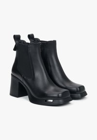 Zwarte leren enkelboots met een blokhak, voorzien van elastische zijpanelen, subtiele stiksels en een metallic logoaccent aan de zijkant.