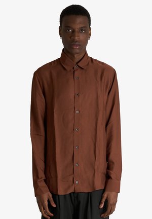 Uomo che indossa una camicia marrone a maniche lunghe con bottoni e colletto, in piedi davanti a uno sfondo bianco semplice.