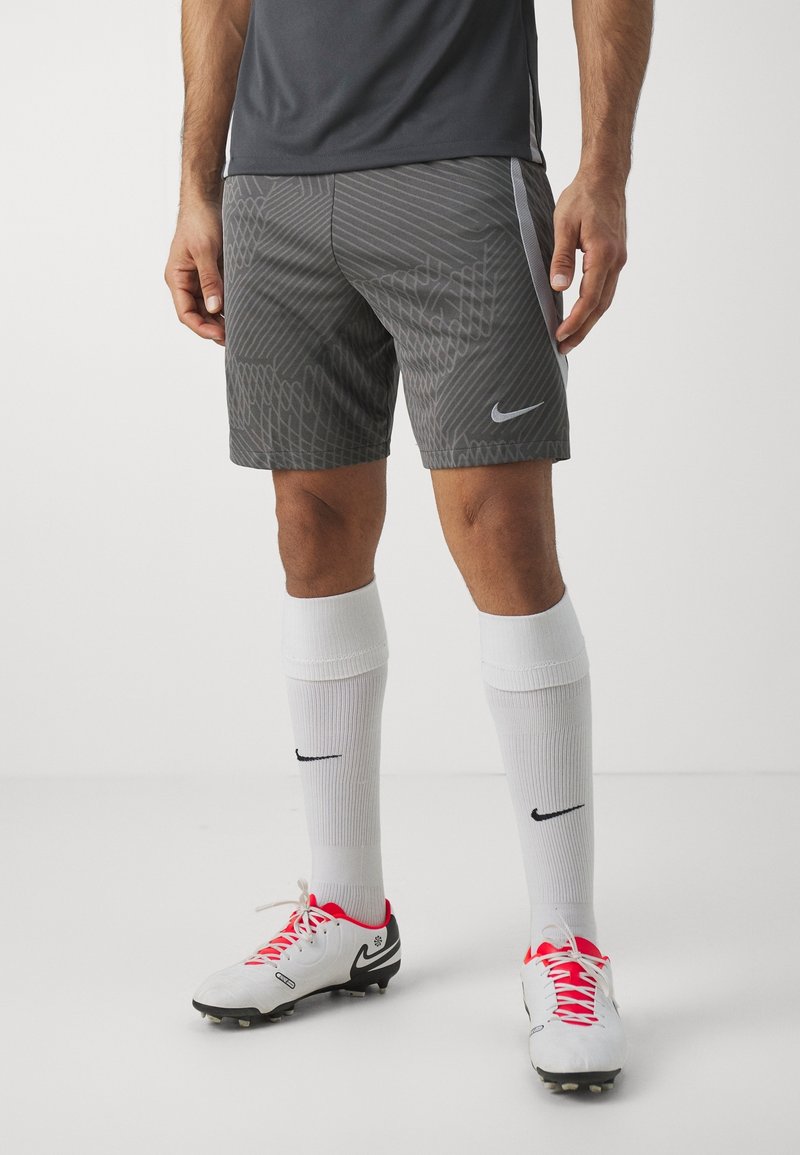 Homme portant un short de sport Nike gris, des chaussettes blanches Nike montant jusqu'aux genoux et des crampons blancs avec des lacets rouges, debout sur un sol blanc devant un fond uni.