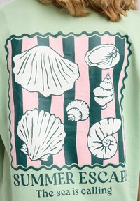 Felpa verde con stampa di conchiglie in bianco e verde scuro su uno sfondo a righe rosa. Il testo recita "EVASIONE ESTIVA" e "Il mare chiama."