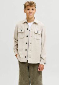 Teenager con i capelli ricci indossa una giacca beige con bottoni e tasche a pattina, una camicia bianca e pantaloni in velluto a coste color oliva, in piedi davanti a uno sfondo neutro.