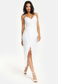 White dress with lace overlay, featuring a fitted bodice, spaghetti straps, and a thigh-high slit. Accented with scalloped edges.