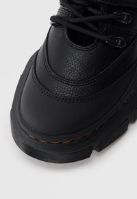 Baskets en cuir noir avec finition texturée, bout rond, semelle en caoutchouc contrastante et lacets noirs. Présente des accents de couture jaunes.