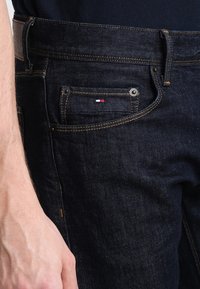 Dark denim jeans featuring orange stitching, a small pocket with a red, white, and blue logo, and metal hardware on the front.