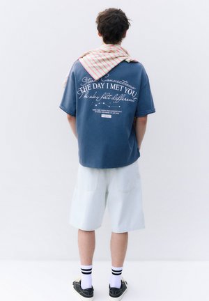 Man draagt een blauw bedrukt T-shirt, witte knielange shorts, zwarte sneakers en een gestreepte doek over zijn schouders, staand tegen een witte achtergrond.
