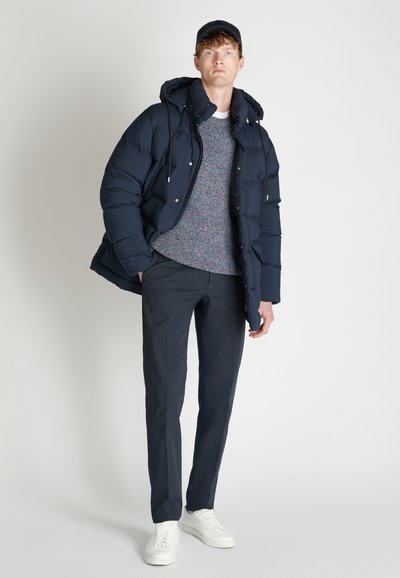 Giacca a vento navy con cappuccio, maglione a maglia testurizzata, pantaloni scuri e sneakers bianche. Modello in piedi con le mani in tasca contro uno sfondo neutro.