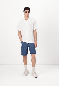Camisa branca de manga curta com um padrão texturizado, combinada com calções de ganga azuis e ténis brancos com detalhes cinza claro.
