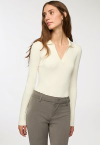 Jasnobiały, długi rękaw, prążkowany body z dekoltem w kształcie litery V i kołnierzykiem polo, noszone z szarymi, dopasowanymi spodniami. Gładka faktura i dopasowany krój.