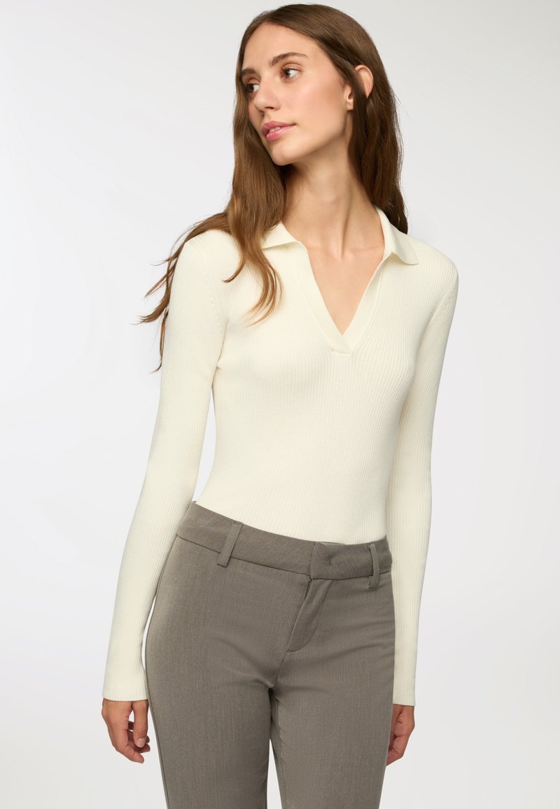 Jasnobiały, długi rękaw, prążkowany body z dekoltem w kształcie litery V i kołnierzykiem polo, noszone z szarymi, dopasowanymi spodniami. Gładka faktura i dopasowany krój.