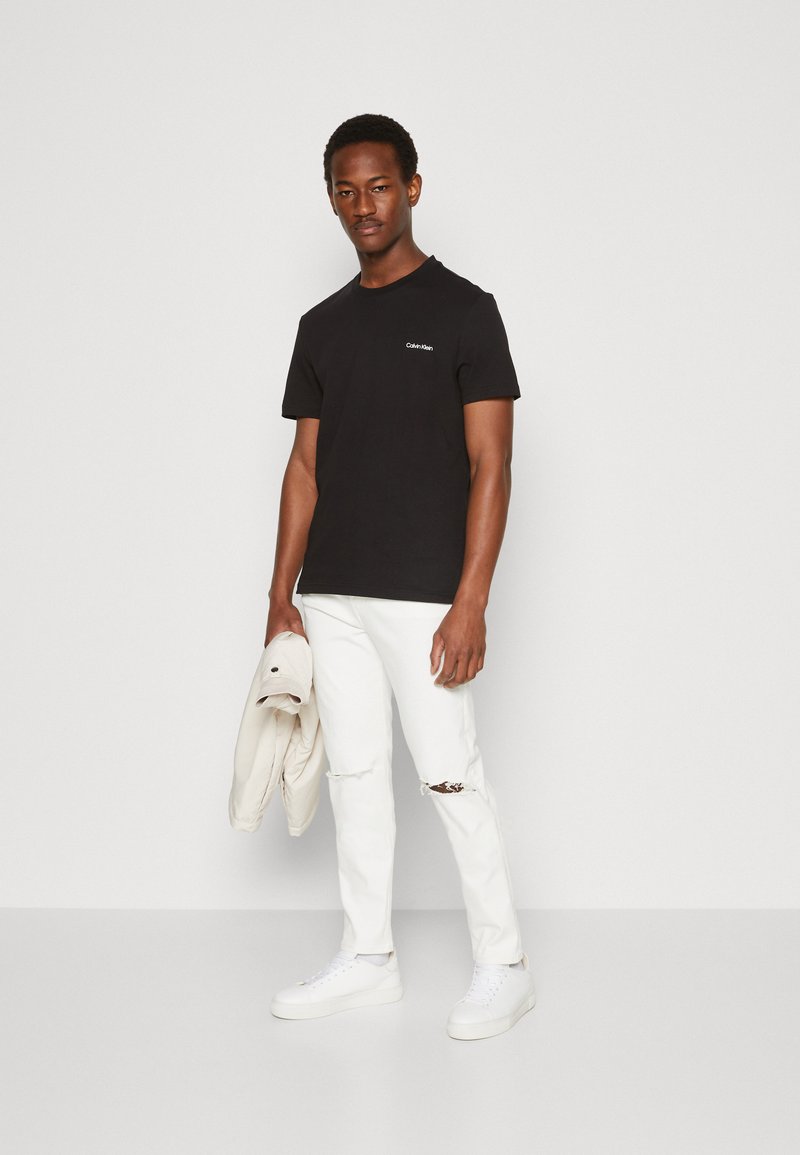 Sort bomulds T-shirt med rund hals og lille hvidt logo, parret med hvide slidte jeans og hvide sneakers. Afslappet outfit.