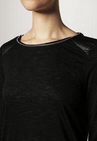 Czarna bluzka z długim rękawem z teksturowanym tricotem, gładkimi czarnymi akcentami na ramionach i zamek błyskawiczny przy dekolcie. Delikatnie opadający materiał.