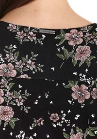 Tela negra con un estampado floral que presenta flores rosas y hojas verdes. Incluye una pequeña etiqueta plateada de la marca en el escote.