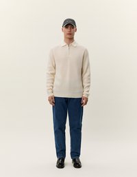Pullover a coste color beige chiaro con colletto a zip, abbinato a jeans in denim blu e scarpe in pelle nere, completato da un cappellino scuro.