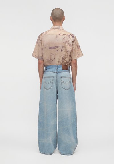 Άτομο με ξυρισμένο κεφάλι που φοράει μπεζ λουλουδάτο πουκάμισο με κοντά μανίκια, μπλουζάκι μαλακό, μέσα σε φαρδιά γαλάζια τζιν παντελόνια, στέκεται γυρισμένο απέναντι.