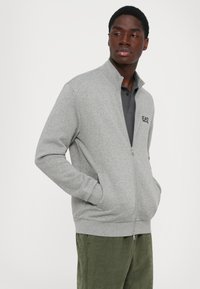 Mann iført en grå zip-up jakke med hendene i lommene, mørk grå polo skjorte under, og grønne bukser, ser til siden.