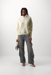 Krema kapuca s prednjim džeparom i tekstualnim detaljima, uparen s svijetlim cargo trapericama koje imaju bočne đepove, drži srebrnu torbicu.