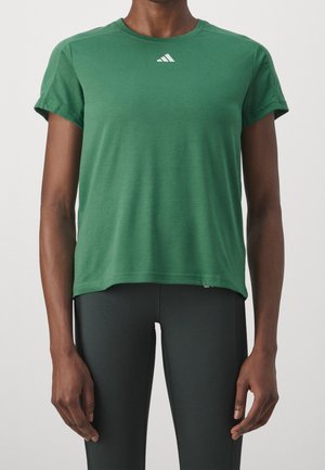 Person, die ein kurzärmeliges grünes Shirt mit kleinem weißem Logo auf der Brust und schwarze enganliegende Hose trägt, steht vor einem einfarbigen Hintergrund.
