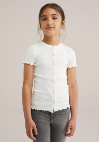 Mlada djevojka s smešnom kosom vezanom unatrag, nosi bijelu košulju na dugmad s kratkim rukavima i sive traperice, stoji naslonjena na jednostavnu pozadinu.