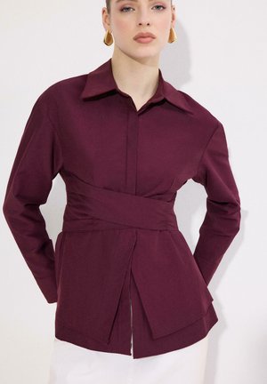 Femme portant un chemisier cache-cœur bordeaux profond avec un large col et des manches longues, associé à un bas blanc et des boucles d'oreilles géométriques dorées.