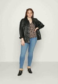 Casaco de pele sintética preto sobre uma blusa de padrão de leopardo, combinado com jeans skiny azul claro e mules de salto pretos. Fundo simples.