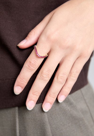 Zlatý prsteň v tvare V s ružovými drahokamami, ktorý má na obvode pruhovaný vzor, nosený na ruke so svetlou pokožkou.