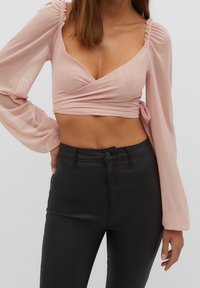 Lichtroze crop top van doorschijnende stof, met een wikkelontwerp, lange pofmouwen en een strikdetail in de taille. Draagt het met zwarte broeken.