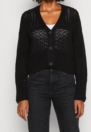 Mujer con un cárdigan de punto crochet negro con botones sobre una camiseta blanca y jeans gris oscuro, vista desde el cuello hasta las caderas.