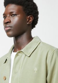 Lichtgroene denimjas met een kraag, voorzien van een knoopsluiting en een klein geborduurd boomlogo op de borst. Zachte textuur.