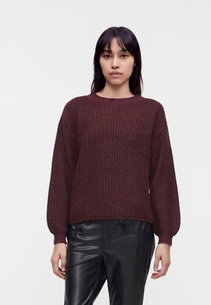 Jeune femme aux cheveux raides foncés porte un pull en maille bordeaux et un pantalon en cuir noir, debout devant un fond clair uni.
