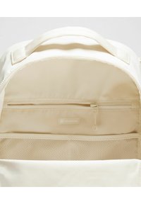 Interior de mochila crema con un bolsillo con cremallera, un compartimento de malla y una etiqueta. El material parece suave con un acabado mate.