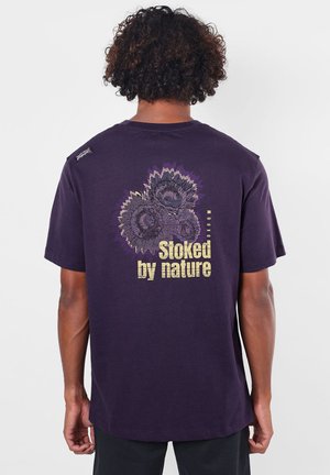 Homme aux cheveux bouclés portant un t-shirt violet foncé avec un motif de tournesol et le texte "stoked by nature" dans le dos.