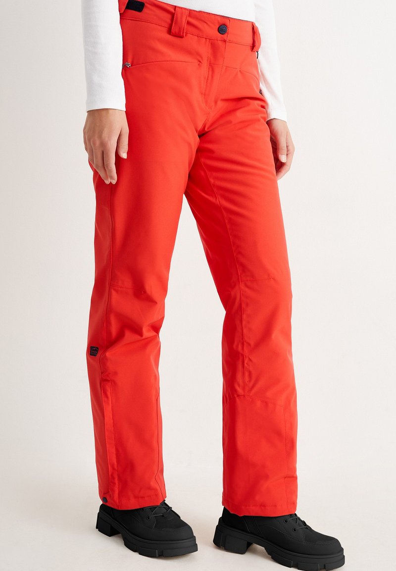 Frau in leuchtend roten Schneehosen mit schwarzen Stiefeln und einem weißen Langarmshirt, steht vor einem weißen Hintergrund.