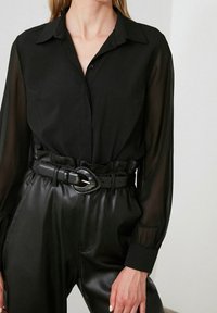 Camicia nera con bottoni e maniche lunghe trasparenti, abbinata a pantaloni in pelle nera a vita alta e una cintura nera larga con fibbia.
