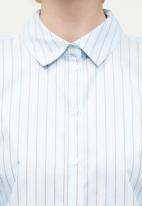 Ανοιχτό μπλε πουκάμισο με λεπτές καφέ κάθετες ρίγες, στρογγυλεμένο γιακά και λευκά κουμπιά μπροστά.