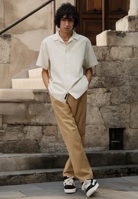 Chemise beige à manches courtes avec boutons, pantalon beige, et baskets noir et blanc. Cheveux bouclés et collier en chaîne léger. Fond en pierre.