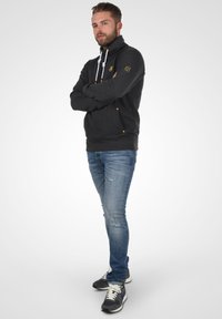Sudadera de forro polar gris oscuro con cuello alto, cordones blancos, acentos amarillos y dos bolsillos frontales, combinada con unos vaqueros desgastados de color azul claro y zapatillas.