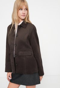 Brown suede jacket with a collar, front buttons, and a chest pocket. Worn with a light top and gray skirt. Smooth texture, modern design.