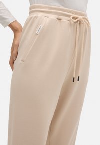 Pantalones deportivos beige con cinturilla elástica, cordón, bolsillo lateral y una pequeña etiqueta blanca de la marca en el lado izquierdo.