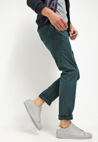 Slim-fit mörkgröna chinos med uppvikta benslut, kombinerade med ljusgrå låga sneakers, med en slät tygtextur.