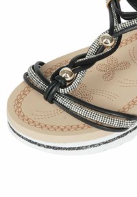 Sandalo nero con cinturini intrecciati, impreziosito da strass argentati e dettagli dorati. Plantare beige testurizzato con motivi decorativi. Suola bianca.