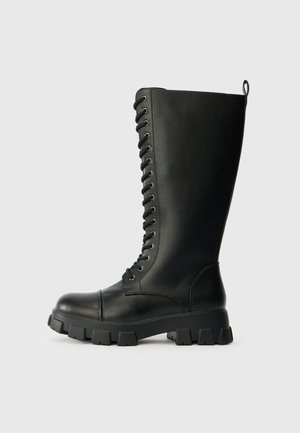Black leather knee-high boots with a sturdy, textured upper, round toe, prominent laces, and chunky, treaded rubber sole.