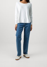 Maglia bianca a maniche lunghe con scollatura tonda, abbinata a jeans blu a gamba dritta e sneakers bianche con suole marroni. Tessuto dalla texture liscia.