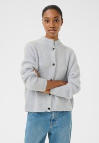 Světle šedý žebrovaný pletený cardigan s vysokým límcem, zapínáním na knoflíky a ohrnutými rukávy, spárovaný s modrými džíny.
