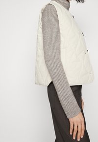 Prošívaná krémová vesta s vysokým límcem a bočními knoflíky, vrstvená přes žebrovaný šedý svetr. Vyznačuje se hladkou, měkkou texturou.