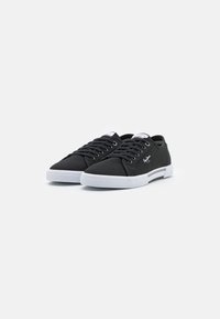 Black canvas sneakers with white rubber soles and laces, featuring a textured toe cap and side logo detail. Classic low-top design.