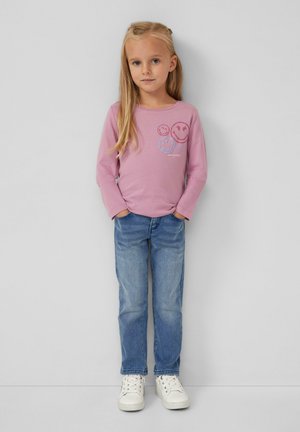 Ragazzina con lunghi capelli biondi che indossa una maglietta rosa a maniche lunghe con faccine sorridenti, jeans blu e sneaker bianche, in piedi con le mani nelle tasche.